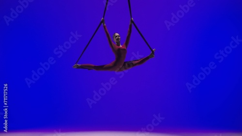 Female gymnast isolated on blue neon studio background. Girl aerial dancer holding and spinning on acrobatic straps in split. © kinomaster