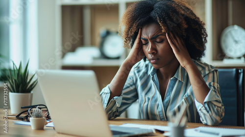 Woman feeling stressed while working on her laptop. She has her head in her hands, a pained expression on her face, signifying a headache, frustration, or exhaustion. photo