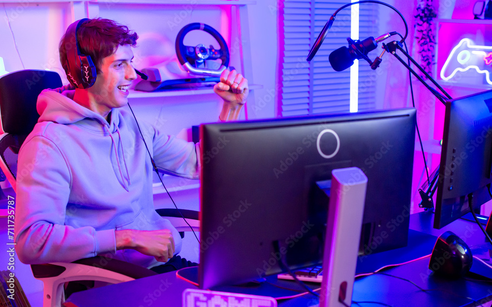 Handsome happy young male game online streamer or caster smiling with fun, raising arms, succeed for winner competition, using computer to play, sitting in room at home with decorated neon light.