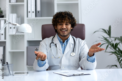 Cheerful male physician in lab coat with stethoscope in an office setting, exhibiting a warm, inviting gesture.