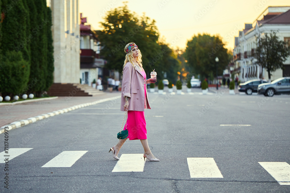 Stylish female blonde walking on a street. Elegant woman wearing trendy pink suit and trousers, sunglasses, with textured green shoulder bag. Girl holding a drink in her hands. Summer photo.