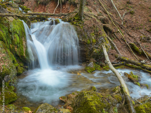 A huge debit waterfall flowing through large mossy stones and boulders, branches and tree trunks, forming a pond. The water is azure colored. Spring season, Sureanu Mountains, Carpathia, Romania 