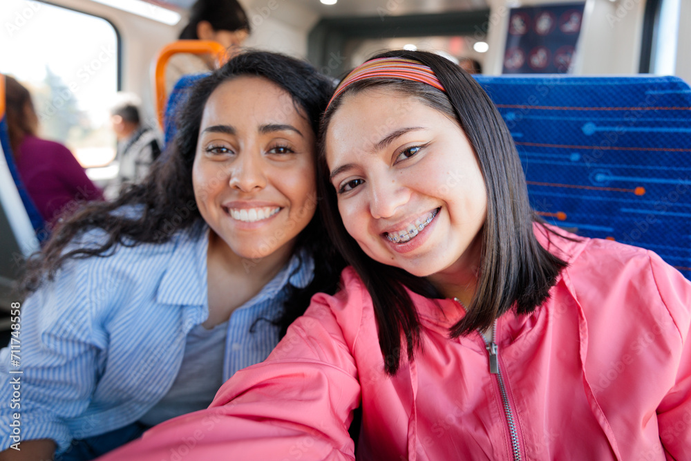 Two young cheerful latin student women traveling on public train transportation in the city and taking selfie with phone Two young cheerful latin student women traveling on public train transportation in the city and taking selfie with phone