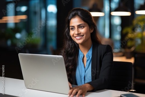 A woman is seen sitting in front of a laptop computer, engaged in online work or study, Happy young indian businesswoman using computer sit at office desk, AI Generated