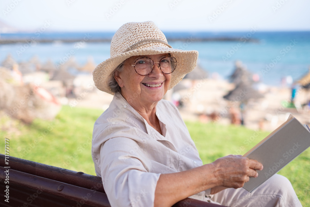 Handsome smiling senior woman sitting outdoor in a bench reading a book enjoying retirement lifestyle. Beach sea and horizon on background