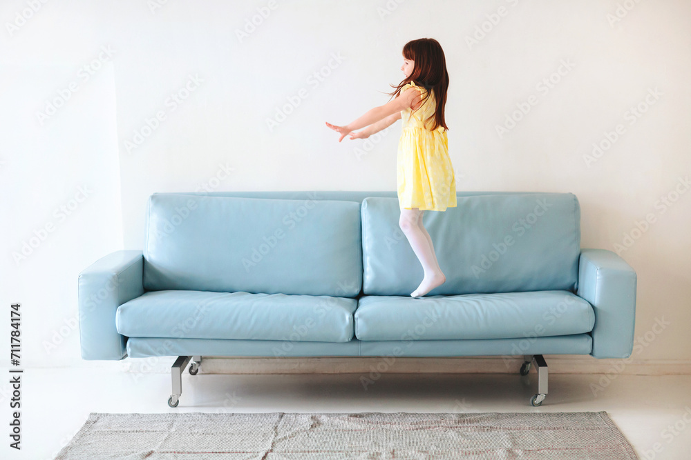 Little caucasian cute girl in yellow dress playing and jumping on blue sofa in living room at home Little caucasian cute girl in yellow dress playing and jumping on blue sofa in living room at home