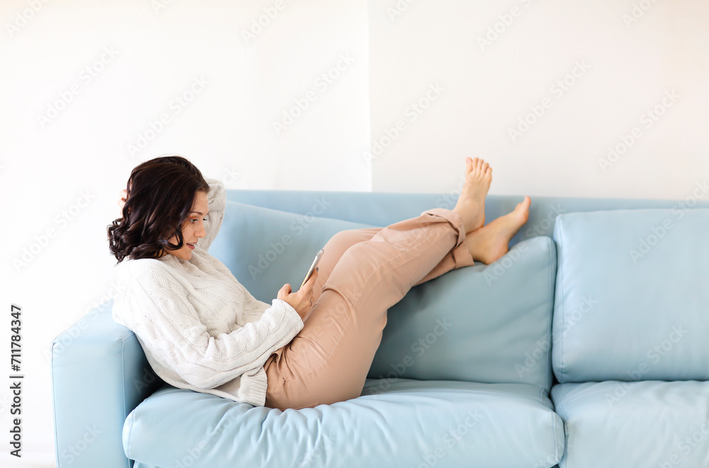 Young brunette woman laying down on cozy sofa in relax position with mobile phone in hands Young brunette woman laying down on cozy sofa in relax position with mobile phone in hands