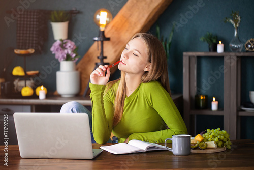 Fotomural Young pensive girl sitting at table at home with laptop, making notes, staring t