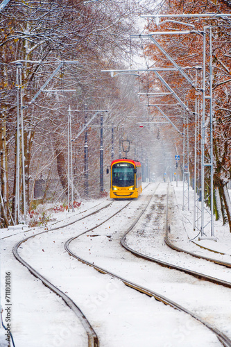 Jadący tramwaj w Warszawie na Bielanach w scenerii zimowej przy padającym śniegu pośród drzew..