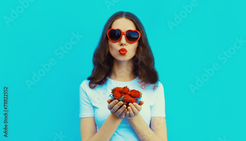 Portrait of beautiful caucasian young woman with handful of fresh strawberries blowing her lips wearing red heart shaped sunglasses on blue studio background