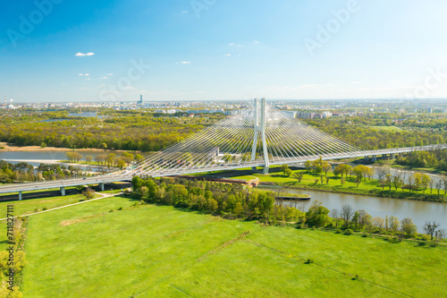 Cars drive on cable-stayed Redzinski Bridge over river flowing near scenic Wroclaw. Pylon bridge surrounded by lush green forests aerial motion along bridge photo