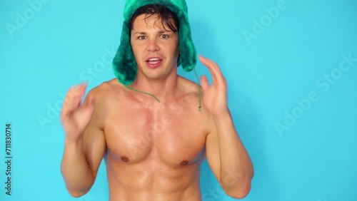 Young sexy man in green fur cap dances and sings in blue studio photo