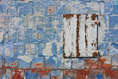 Old wall with peeling paint and closed white window. Colored background of house with scratched blue plaster.