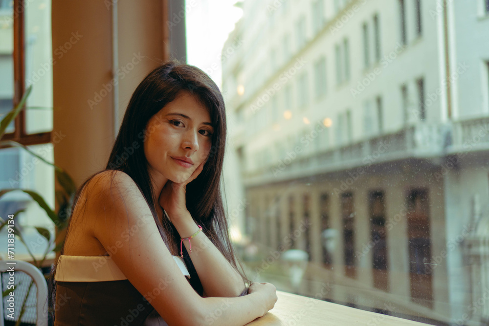 young Latin woman looks at the camera satisfied, sitting in a café with a view of the city. copy space