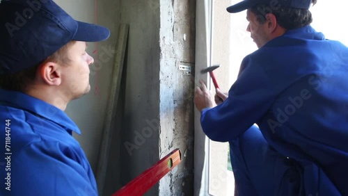 Worker hammers dowel fixing bracket on jamb. Another one stands near photo