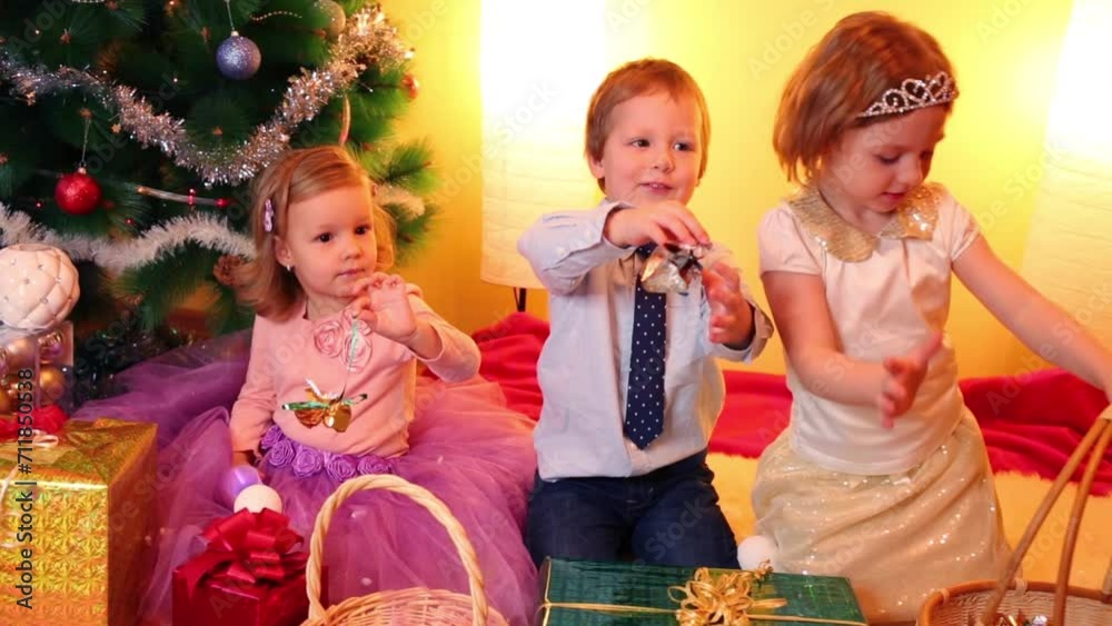 Three children sit in room under christmas tree among gift boxes Three children sit in room under christmas tree among gift boxes
