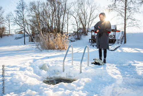 Man in winter gear standing by a swimming hole cut in the snow, with a serene winter landscape around