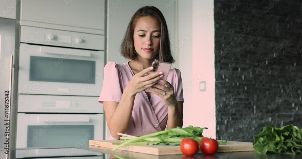 Young attractive female holding modern smart phone, distracted from healthy vegetable salad preparation. Food blogging, on-line cookbook recipes application usage, chatting while cooking in kitchen