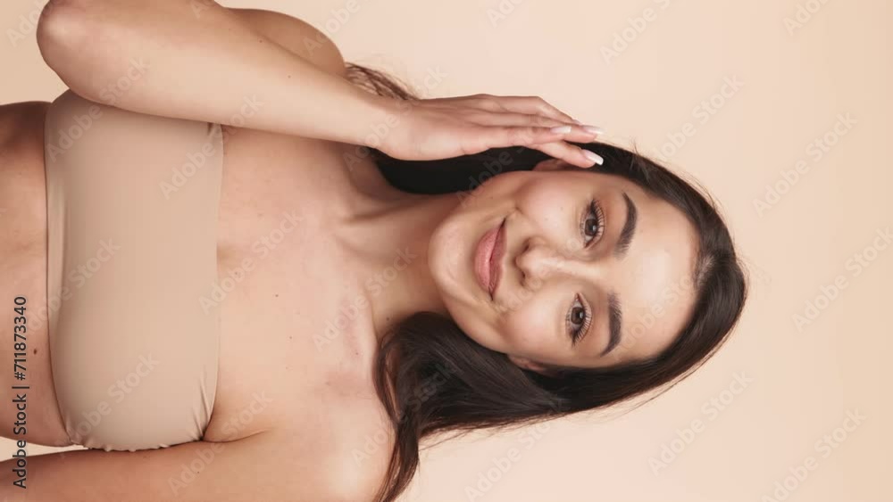 Vertical shot of a graceful young woman showcasing her natural complexion and beauty with a gentle touch to her face against a neutral background.