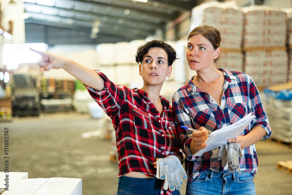 Portrait of caucasian and latin women working in warehouse, discussing job, checking documentation and pointing finger.