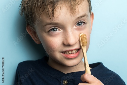 Smiling 6-year-old boy holding a bamboo toothbrush against a light blue background. Eco-friendly dental care concept. Bright portrait promoting sustainable hygiene habits for kids.