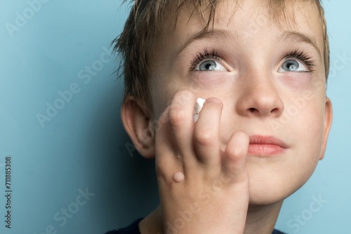 Close-up portrait of a 6-year-old boy applying white cream to his cheek. His sparkling eyes and unique detail of a wart on his hand stand out against a soft blue background