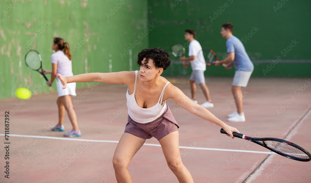 Active fit young hispanic woman playing frontenis on open court on summer day, hitting ball with strung tennis racquet to score to opposing team. Popular Spanish sports..