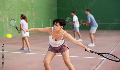 Active fit young hispanic woman playing frontenis on open court on summer day, hitting ball with strung tennis racquet to score to opposing team. Popular Spanish sports..
