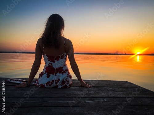 Rear view of woman sitting down on a wooden pontoon near the ocean and admiring the sunset