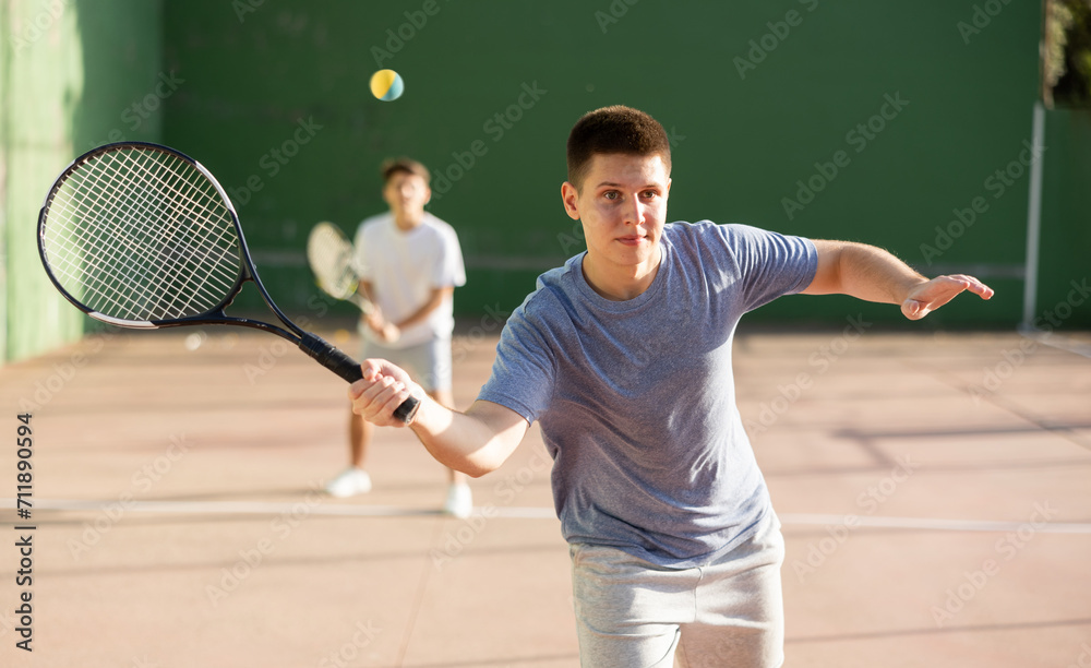 Emotional athletic young guy playing frontenis on open court on summer day, hitting ball with strung tennis racquet to score to opposing team