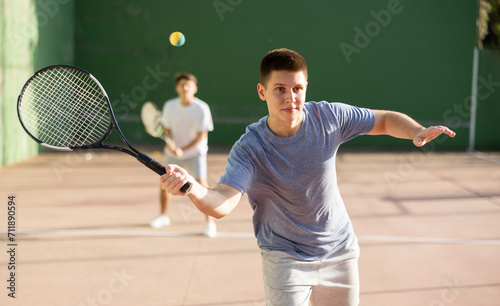 Emotional athletic young guy playing frontenis on open court on summer day, hitting ball with strung tennis racquet to score to opposing team