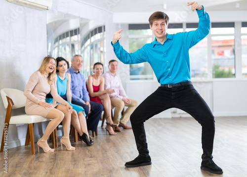 Expressive cheerful young guy wearing black jeans and blue shirt demonstrating krump dance moves to mix-aged group of amateurs in modern choreographic studio.. photo