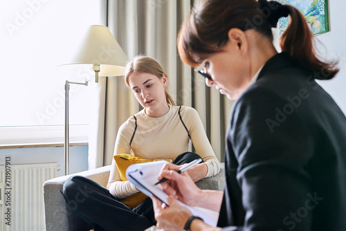 Young female teenager talking with counselor, teacher sitting in chair in office