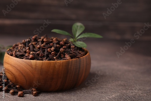 Aromatic cloves and green leaves in bowl on brown table, closeup. Space for text