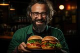 A bearded man indulges in a mouthwatering american fast food snack, holding a plate of juicy burgers with glasses perched on his face in an indoor setting