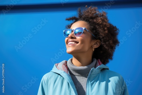 Young girl standing in blue background wearing a blue hoodie and sunglass