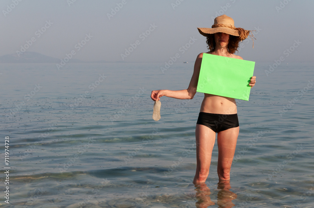 Mid Adult Woman in Swimsuit and Straw Hat on a Beach holding over Breasts a Green Screen Canvas and in other hand Hands a piece of Garbage found in sea