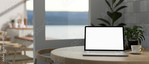 A white-screen laptop computer mockup and a coffee cup on a table in a beautiful modern coffee shop. © bongkarn