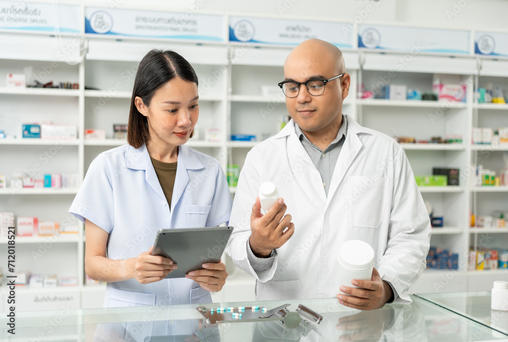 Two asian man and woman professional Pharmacist colleagues working at drugstore pharmacy. Asking the questions of medication standing near pharmacy shelves counter with medicines Two asian man and woman professional Pharmacist colleagues working at drugstore pharmacy. Asking the questions of medication standing near pharmacy shelves counter with medicines