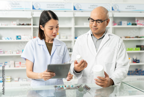 Two asian man and woman professional Pharmacist colleagues working at drugstore pharmacy. Asking the questions of medication standing near pharmacy shelves counter with medicines