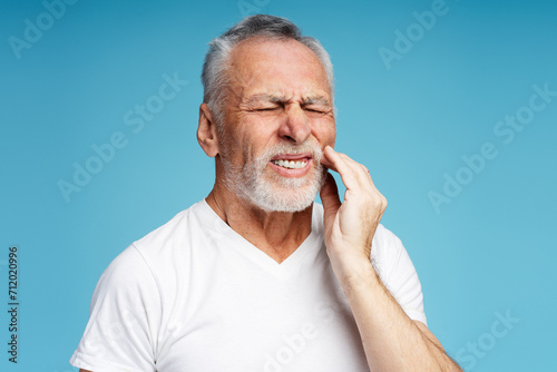 Upset mature retired man wearing white casual t shirt with toothache isolated on blue background photo