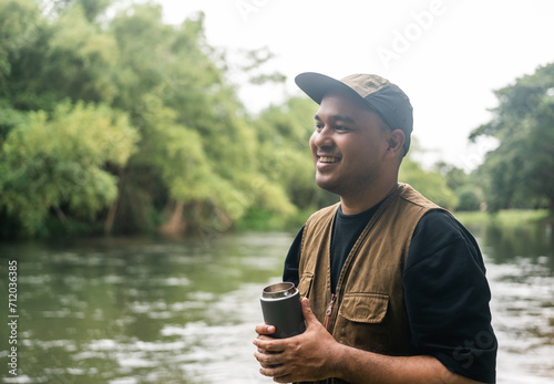 Young asian man traveller relaxing drinking coffee in nature forest and river background. Happy male living in middle of a lot of tree. Breathing the fresh air on her vacation weekend holiday trip.