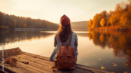 traveler with backpack relaxing by autumn river at sunset. young woman raised arms feeling free and happy