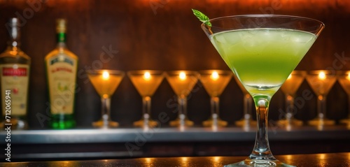  a green drink in a martini glass with a sprig of mint in front of a row of champagne glasses on a bar with liquor bottles in the background.