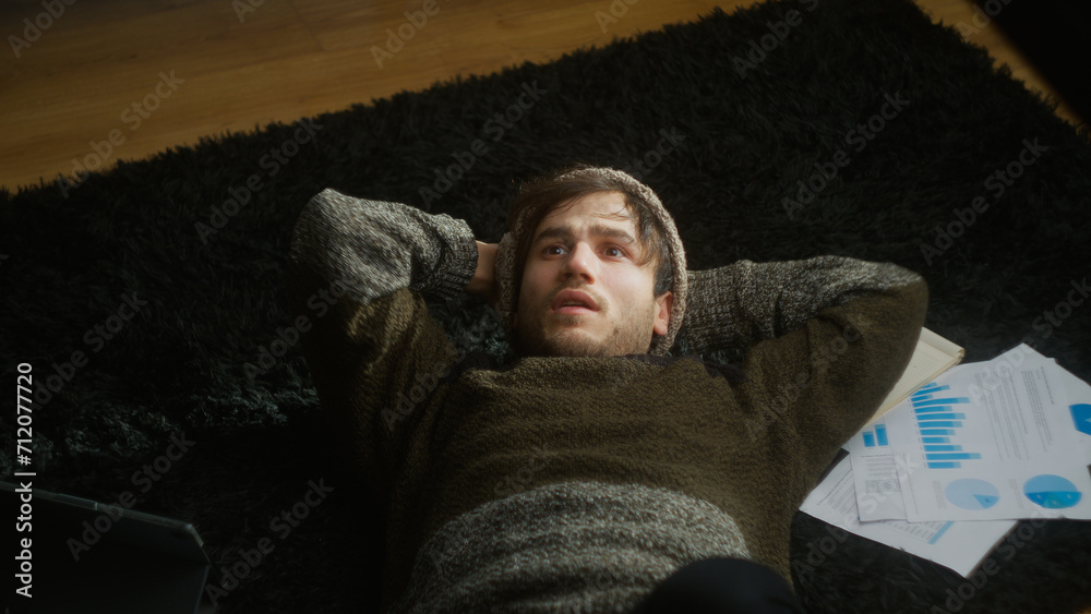 Fototapeta premium Thoughtful young student with beanie, lying on the carpet on the floor, comes up with a great idea, picks up one of the notes on the floor and overjoy 