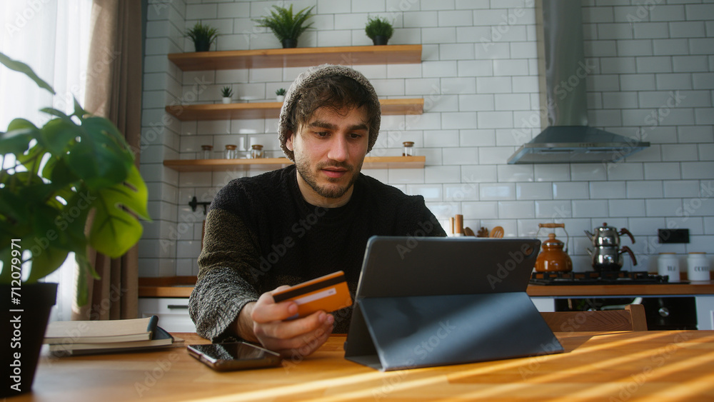 Man with beanie sitting in kitchen at home entering credit card number on tablet for makes secure easy distant electronic payment Man with beanie sitting in kitchen at home entering credit card number on tablet for makes secure easy distant electronic payment