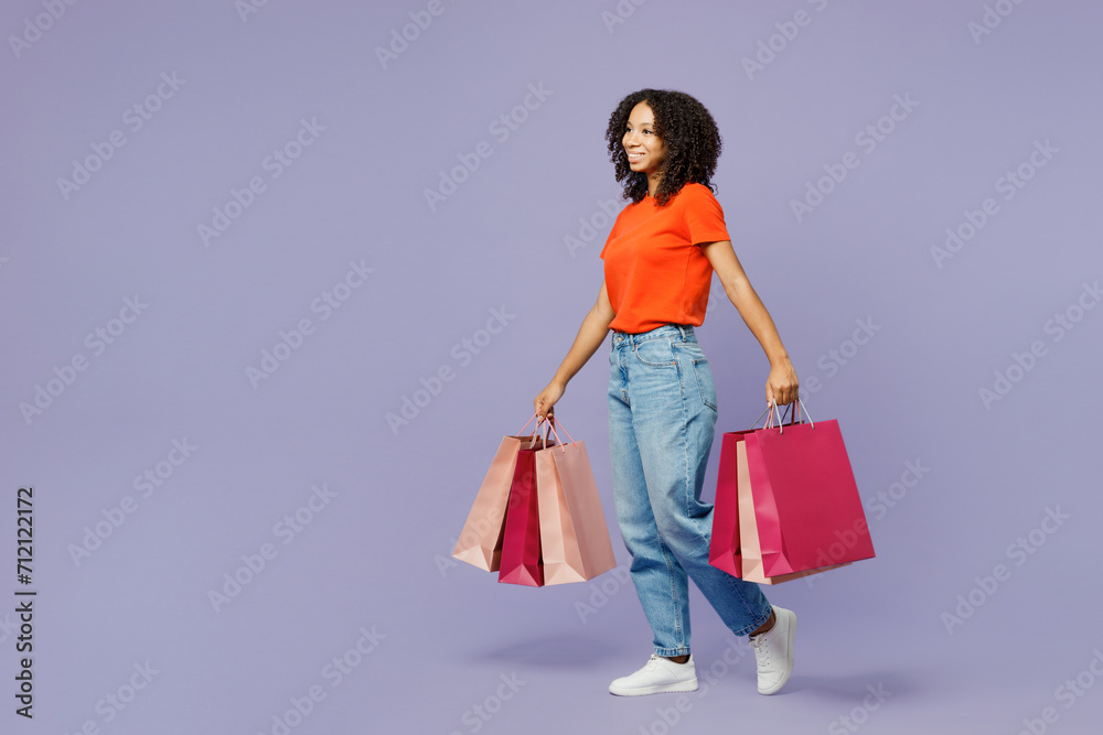 Full body little kid teen girl of African American ethnicity wear orange t-shirt hold paper package bags after shopping isolated on plain purple background studio. Black Friday sale buy day concept.