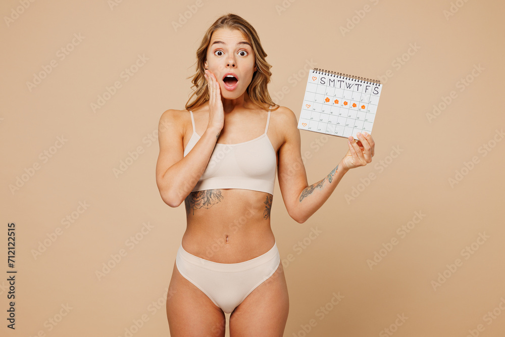 Naklejka premium Young shocked nice lady woman with slim body perfect skin wear nude top bra lingerie stand hold checking menstruation days calendar isolated on plain beige background. Lifestyle gynecological concept.