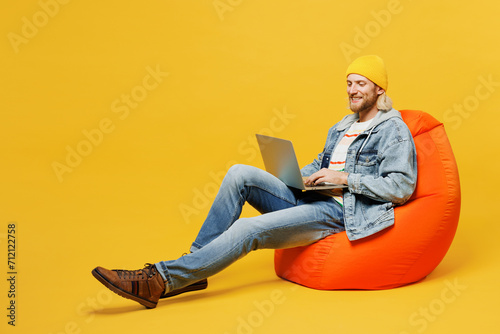 Full body young IT man he wears denim shirt hoody beanie hat casual clothes sit in bag chair hold use work on laptop pc computer isolated on plain yellow background studio portrait. Lifestyle concept.