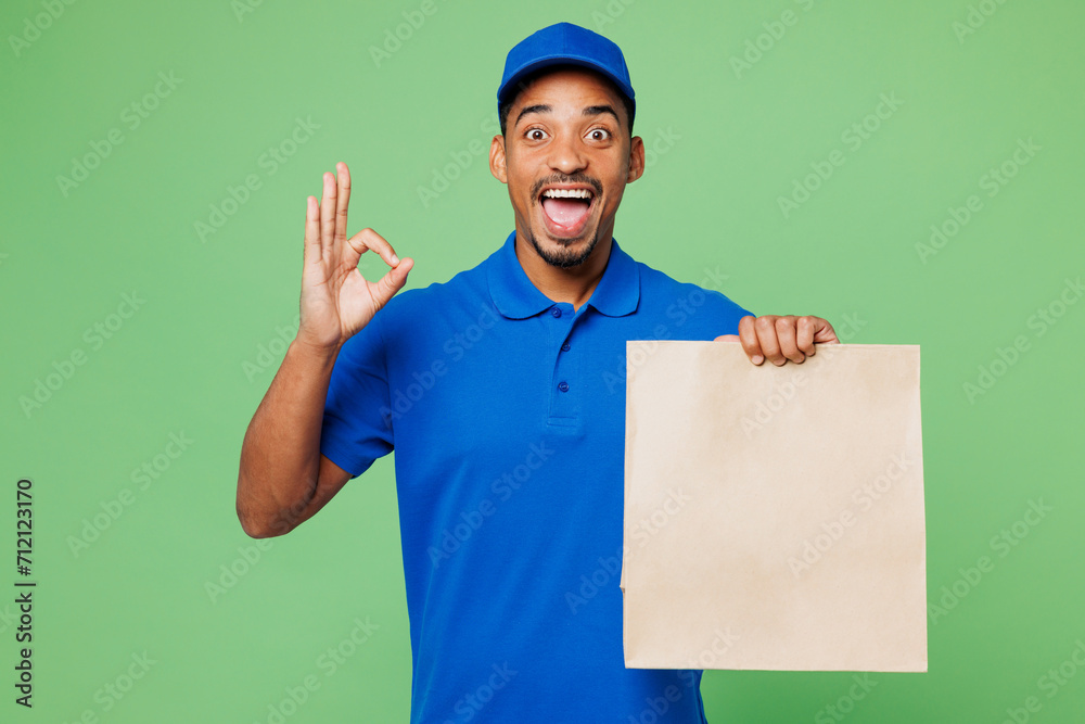 Delivery guy employee man wears blue cap t-shirt uniform workwear work as dealer courier hold blank craft paper takeaway bag mock up show ok okay isolated on plain green background. Service concept. Delivery guy employee man wears blue cap t-shirt uniform workwear work as dealer courier hold blank craft paper takeaway bag mock up show ok okay isolated on plain green background. Service concept.